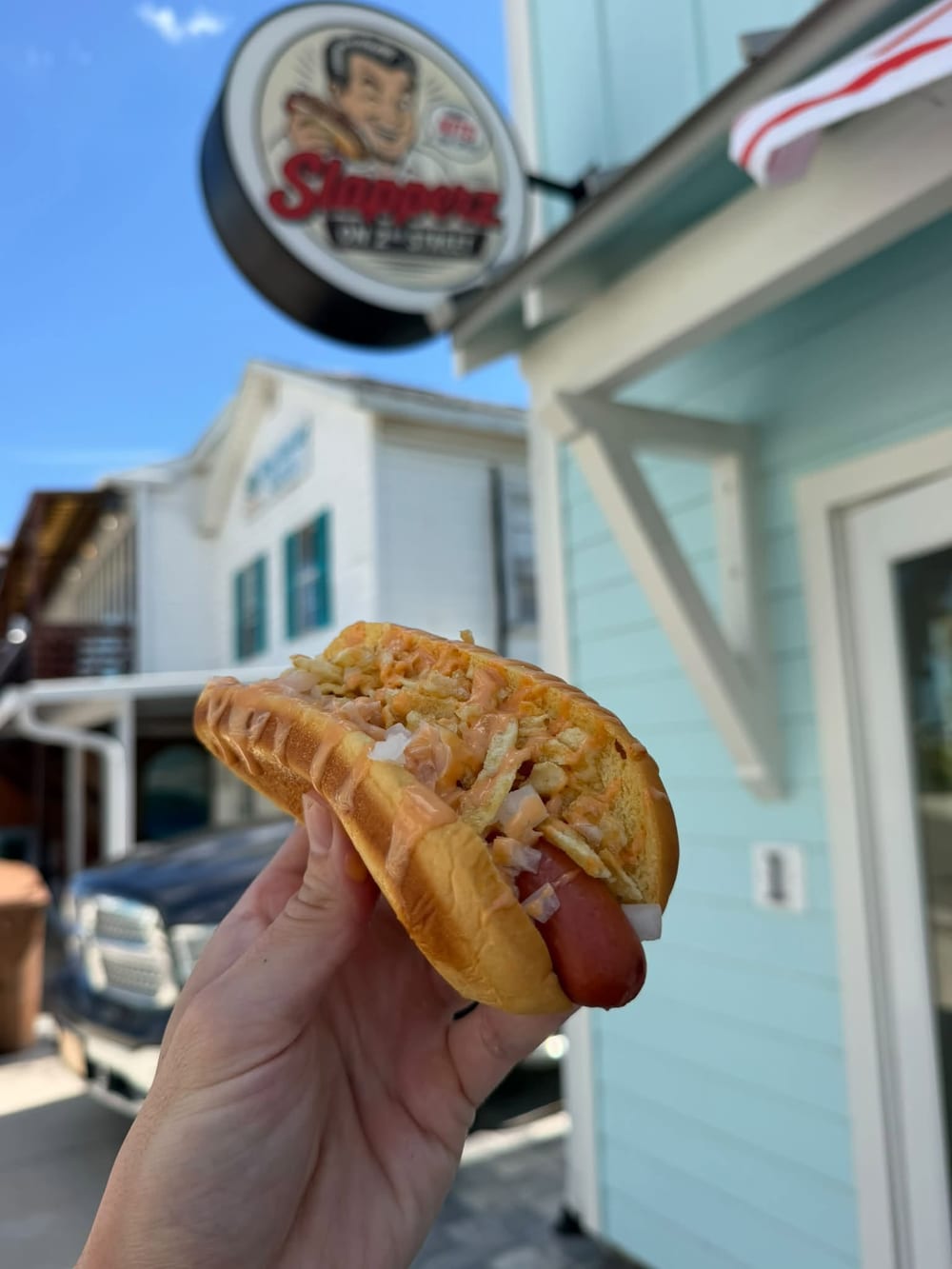 A Slapperz Signature Dog on a toasted bun topped with crushed potato chips, chopped white onion and orange sauce, held up in front of the Slapperz sign
